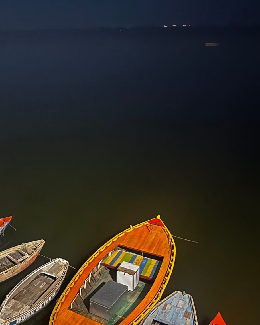 Wooden boats moored on the Ganga at night, with distant lights along the far bank